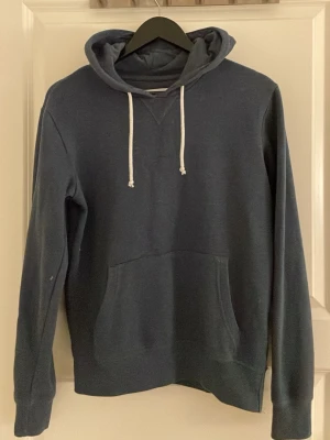 Mörkblå hoodie med ficka och huva - En stilren mörkblå hoodie med klassisk känguruficka framtill och vit dragsko i huvan. Hoodien har långa ärmar och ribbade muddar vid ärmslut och nederkant. Perfekt för en avslappnad look och enkel att matcha med jeans eller joggers.