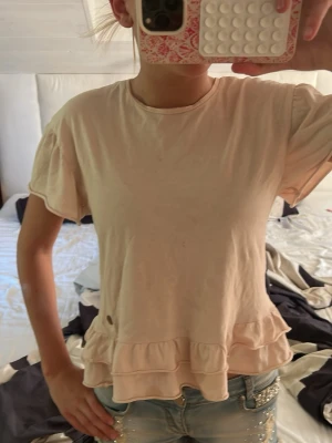 Ljusbeige topp med volangkant - Söt ljusbeige topp med korta ärmar och dubbla volangdetaljer längst ner. Tillverkad i mjuk bomull och har en avslappnad passform. Perfekt att styla med jeans eller kjol för en trendig look. Storlek 146-152 så ca xxs, den är från Ü lite fläckar järndörrarna har aldrig testat att ta bort dem förutom att ha tvättat tröjan jag kan inte lova att dem går bort!