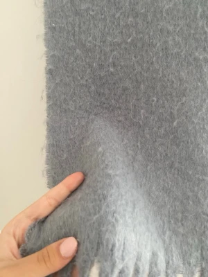 Ljusgrå fluffig halsduk med fransar - Mysig ljusgrå halsduk i fluffigt material med långa fransar i ändarna. Perfekt för att hålla värmen under kyliga dagar och ger en trendig touch till din outfit. Enkel att matcha med olika stilar och färger.