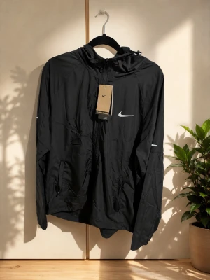 Svart Nike vindjacka med huva - Svart vindjacka från Nike med vit logga på bröstet och reflekterande detaljer på ärmarna. Jackan har dragkedja framtill, två fickor med dragkedja och justerbar huva. Perfekt för blåsiga dagar och sportiga outfits.  Nike seal bag kommer även med 