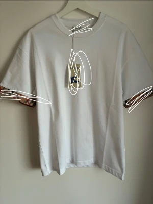 Vit Burberry t-shirt med rutiga detaljer - Vit tröja med burberry märket på sidan av armen. Väldigt bra och trevlig och fräsch nu inför sommaren om man letar efter något som sticker ut. Storlek M men är lite stor i storlek. Priset går att diskutera 