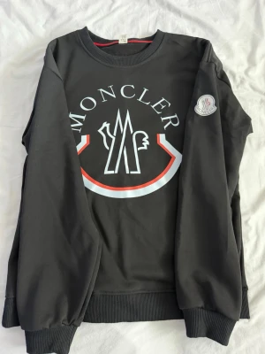Svart sweatshirt från Moncler - Svart sweatshirt från Moncler med stort vitt och rött logotryck på bröstet och Moncler-märke på ärmen. Tröjan har rund hals, ribbade muddar och är gjord i mjukt bomullsmaterial. Perfekt för dig som gillar streetwear och vill ha något stilrent men ändå med tydlig branding.