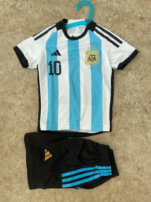 Argentina Messi matchställ - Säljer ett Argentina matchställ från Adidas med Messi och nummer 10. Tröjan är vit och ljusblå med svarta detaljer och AFA-logga på bröstet. Shortsen är svarta med blå ränder och guldfärgad Adidas-logga. Perfekt för fotbollsträning eller att heja på favoritlaget.