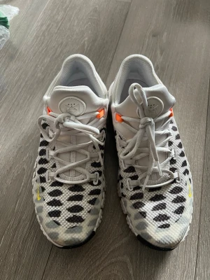 Nike vita sneakers med svart mönster - Säljer ett par unika vita Nike kängor med svart prickigt mönster och coola detaljer i orange och gult. Skorna har en chunky sula och blå detalj på hälen. Tillverkade i mesh och syntetmaterial för en sportig look. Perfekta för dig som vill sticka ut. Storlek är 40 men passar för 38.39 och 40. 