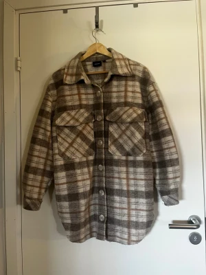 Rutig overshirt från Gina Tricot - Säljer en oversized rutig overshirt från Gina Tricot i beige, brunt och vitt. Skjortjackan har stora bröstfickor med lock, knäppning framtill och klassisk krage. Tillverkad i mjukt, borstat material som ger en chill och trendig look.
