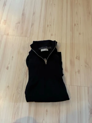 Svart tunn tröja från Jack & Jones - Snygg svart tröja från Jack & Jones med halv dragkedja framtill. Klassisk design med hög krage och mjukt material som känns skönt mot huden. Perfekt för dig som gillar en enkel och stilren look. Passar till det mesta och är lätt att matcha.