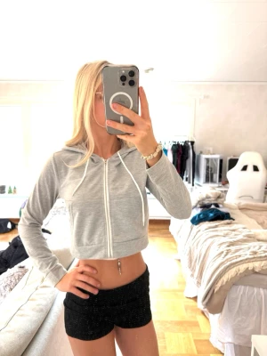 Ljusgrå cropped hoodie med dragkedja - Snygg ljusgrå cropped hoodie med dragkedja framtill och huva med dragsko. Hoodien har långa ärmar och ribbade muddar. Perfekt till jeans eller shorts för en avslappnad look. 