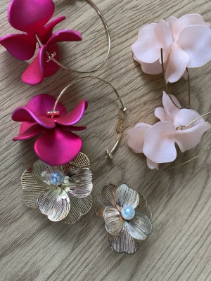 Blommiga örhängen i guld och rosa - Två par stora örhängen med blommor. Ett par har guldiga metallblommor med pärldetalj i mitten, det andra paret har stora rosa och ceriserosa blommor i plast på guldfärgade ringar. Perfekta för dig som vill sticka ut med färg och form.