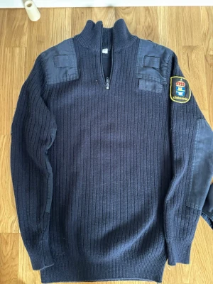 Mörkblå stickad tröja Kriminalvården - Mörkblå stickad tröja med halv dragkedja och ribbad struktur. Tröjan har förstärkningar på axlar och armbågar samt ett Kriminalvården-märke på ena ärmen. Långärmad och med hög krage, perfekt för kyligare dagar.