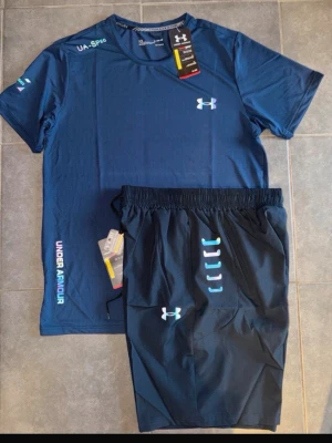 Blått träningsset från Under Armour - Snyggt träningsset från Under Armour med en blå t-shirt och matchande shorts. T-shirten har logga på bröstet och tryck på ärmen samt sidan. Shortsen har elastisk midja, logga och coola detaljer i ljusblått. Perfekt för gymmet eller löpning.