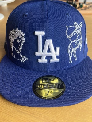 LA Dodgers MLB Mythical Dark Blue 59FIFTY FItted Cap - Snygg blå 59FIFTY keps från New Era med LA-logga framtill. Kepsen har vita broderade motiv av statyer, centaur och pilbåge samt detaljer som årtalet 1920 på sidan. Klassisk flat peak och broderade ventilationshål. Perfekt för dig som gillar streetwear och unika detaljer. Var för liten och har testat den en gång. Helt ny.