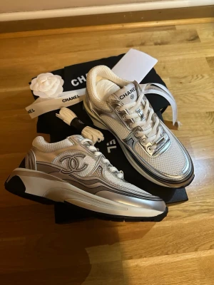 Chanel silvriga sneakers - Chanel sneakers i silver och vitt med mesh-ovandel, metallicdetaljer och tydlig logga på sidan. Skorna har chunky sula i svart och vitt samt snörning framtill. Snygg futuristisk vibe och exklusiv känsla, perfekta för dig som vill sticka ut.