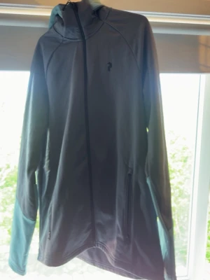  Peak Performance tröja  - Snygg grå vindjacka från Peak Performance med svarta detaljer och ljusare partier på ärmarna. Jackan har huva med logga, hel dragkedja framtill och två fickor med dragkedja. Perfekt för blåsiga dagar och sportiga outfits.