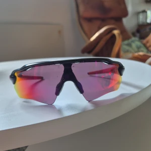 Oakley sportiga solglasögon röd/svart - Säljer ett par sportiga Oakley solglasögon med svart båge och spegellins i rött, rosa och orange. Halvbågsdesign och ikonisk Oakley-logga på sidan. Perfekta för träning och utomhusaktiviteter under soliga dagar.