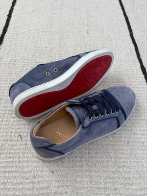 Blåa Louboutin sneakers i mocka - Snygga blåa sneakers från Christian Louboutin i mjuk mocka med klassisk röd sula. Skorna har vita sulor, mörkblå snören och silverfärgade öljetter. Perfekta för dig som vill ha en stilren och exklusiv look.
