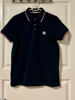 Svart Moncler pikétröja med röd/vit kant - Svart pikétröja från Moncler med korta ärmar och klassisk krage. Kragen och ärmsluten har röda och vita ränder. Tröjan har knappar framtill och Moncler-logga på bröstet. Tillverkad i bomull för en skön känsla.