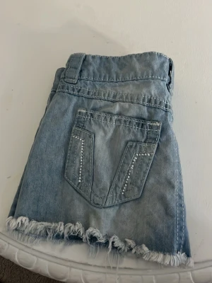 Ljusblå jeansskjol med slitningar - Säljer ett par ljusblå jeansshorts med fransig kant och slitna detaljer. Shortsen har bakficka med dekorativa sömmar och små nitar. Perfekta för sommaren och ger en avslappnad look.