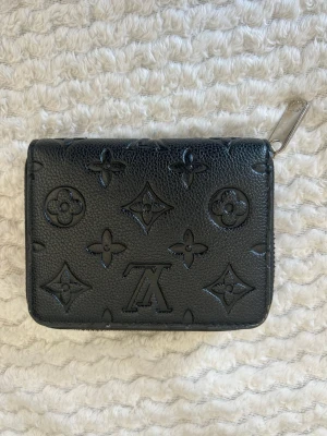 Svart mönstrad plånbok  - Stilren svart plånbok i imitationsläder med monogrammönster. Plånboken har silverfärgad dragkedja och flera fack för kort och sedlar. 