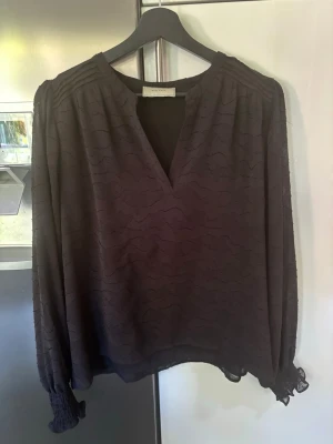 Neo Noir Svart blus med v-ringning och volang - Snygg svart blus från Neo Noir med v-ringad hals och subtilt vågigt mönster i tyget. Långa ärmar med smockade muddar och lätt volangdetalj vid ärmslut. Luftig och flowy passform som ger en avslappnad men chic vibe.