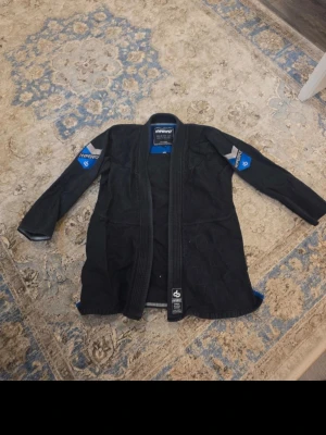 Svart gi-jacka från Hyperfly - Säljer en svart brasiliansk jiu-jitsu gi-jacka från Hyperfly med blå och grå detaljer på ärmarna. Insidan har trycket 'You can't teach heart.' Jackan är tillverkad i slitstarkt bomullsmaterial och har förstärkta sömmar för extra hållbarhet. Perfekt för träning och tävling.