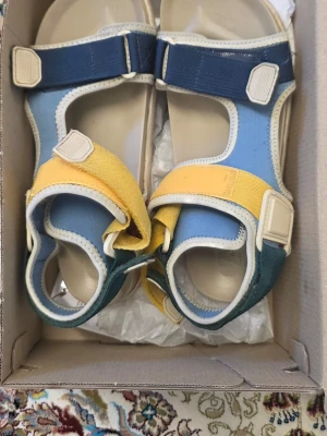 Färgglada sandaler med kardborreband - Säljer ett par sandaler i blått, gult och grönt med vita detaljer. De har justerbara kardborreband och en bekväm sula i beige. Perfekta för sommaren och riktigt snygga till jeans eller shorts. Materialet är textil och syntet.