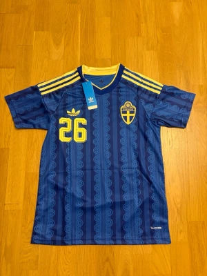 Sveriges vm tröja Taha Ali S - Säljer en blå matchtröja från Adidas med Sveriges landslagsemblem, gula detaljer och nummer 26 på både fram- och baksidan. Tröjan har korta ärmar, gula ränder på axlarna och trycket 'ALI' på ryggen. Tillverkad i lätt funktionsmaterial.