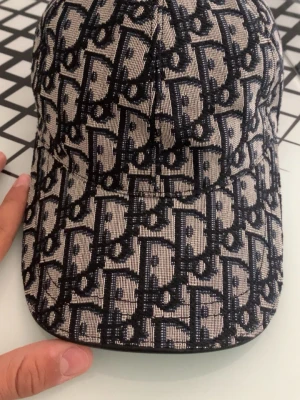 Oblique beige och marinblå keps - Säljer en snygg keps med det klassiska Oblique-mönstret i beige och marinblått. Kepsen har böjd skärm och är tillverkad i ett tygmaterial med broderad logga över hela ytan. Insidan är fodrad i brunt tyg för extra komfort.