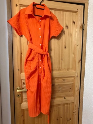 Orange skjortklänning med bälte - Snygg orange skjortklänning med knappar framtill och tillhörande bälte i midjan. Klänningen har korta ärmar, krage och ett lätt ribbat tyg som ger en cool struktur. 