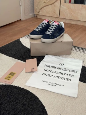 Marinblåa golden goose - Snygga blå sneakers från Golden Goose med vit sula, vita skosnören och vit häl. Skorna har en ikonisk stjärndetalj på sidan och är gjorda i mocka. Perfekta för dig som vill ha en stilren och trendig look.