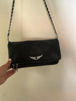  Zadig & voltarie  -  Vet på riktigt itne om den e äkta elr fejk!!!!!!!! Snygg svart axelväska i fejkläder med silverfärgad kedjerem och dragkedjedetaljer. Väskan har ett emblem med vingar framtill och en extra ficka med dragkedja på baksidan. Perfekt storlek för det viktigaste och ger en cool vibe till din outfit.