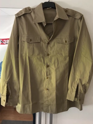 Grön overshirt - Stilren beige skjorta med klassisk krage, axelklaffar och två bröstfickor med knappar. Skjortan har långa ärmar och knäppning framtill. Perfekt för en avslappnad men ändå snygg look.