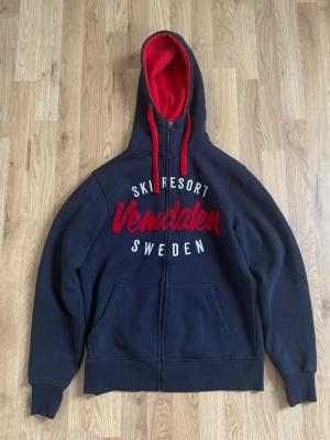 Mörkblå Vemdalen hoodie med dragkedja - Säljer en mörkblå hoodie från United By Nature med röd insida i huvan och röda snören. På bröstet står det 'Ski Resort Vemdalen Sweden' i vitt och rött. Hoodien har dragkedja, kängurufickor och är riktigt mjuk och skön. Perfekt för dig som gillar skidåkning och vill ha en snygg hoodie.