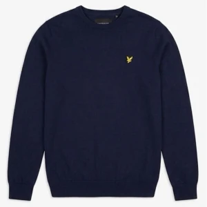 Mörkblå stickad tröja Lyle & Scott - Mörkblå stickad tröja från Lyle & Scott med rund halsringning och den klassiska gula logon broderad på bröstet. Tröjan har långa ärmar och ribbade muddar vid ärmslut och nederkant. Perfekt för lager på lager och enkel att matcha.
