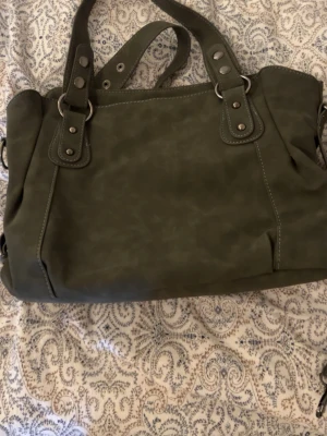 Mörkgrön handväska med nitar - Snygg mörkgrön handväska i fejkläder med dubbla handtag och silverfärgade nitar. Väskan har en mjuk, rektangulär form och dekorativa sömmar. Perfekt för dig som vill ha en stilren och rymlig accessoar.