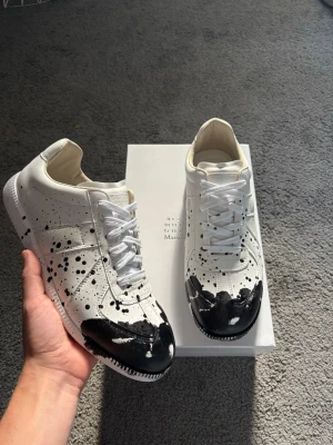 Maison Margiela svartvit sneakers - Unika sneakers från Maison Margiela i vitt läder med svart färgstänk och målad tå. Klassisk låg modell med vita skosnören och vit sula. Perfekt för dig som gillar statement-plagg och konstnärlig stil.