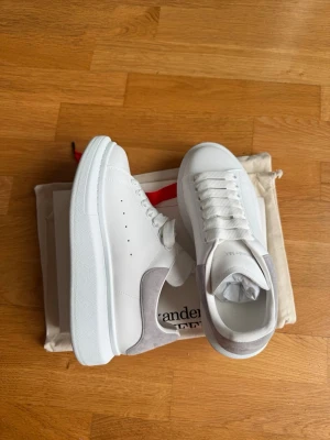 Vita sneakers Alexander McQueen - Stilrena vita sneakers från Alexander McQueen med tjock sula och grå mockadetalj på hälen. Klassisk låg modell med perforerade sidor och vita skosnören. Perfekta för dig som vill ha en clean och trendig look. Bud accepteras men inga skambud då skorna är helt nya. 