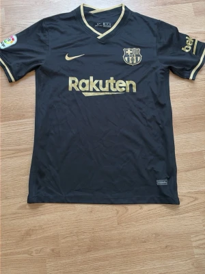  FC Barcelona tröja - Snygg svart FC Barcelona matchtröja från Nike med guldfärgade detaljer och loggor. Rakuten och klubbmärket syns framtill, La Liga-märke på ärmen och UNICEF-tryck baktill. Tillverkad i lätt och ventilerande material, perfekt för fotboll eller träning.