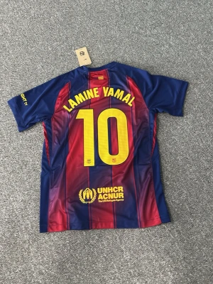 FC Barcelona matchtröja Lamine Yamal - Snygg FC Barcelona matchtröja från Nike med Lamine Yamal och nummer 10 på ryggen. Tröjan har klassiska blå och röda ränder, gula detaljer och klubbmärke på bröstet. Tillverkad i lätt och ventilerande polyester, perfekt för fotboll eller träning.