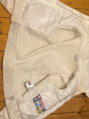 Moncler Cardigan - Snygg vit pufferjacka från Moncler med quiltade partier framtill och på ärmen. Jackan har dragkedja, ribbade muddar och två fickor med knappstängning. Perfekt för kalla dagar och har en sportig, clean look.