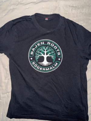 Svart Bajen Roots t-shirt - Snygg svart t-shirt från Bajen Roots med ett stort tryck på bröstet som visar ett vitt träd med gröna blad och texten 'Bajen Roots Södermalm' runtom. Jätte fint skick, använd endast 2 gånger. Storlek L men sitter som M.  Perfekt för dig som vill visa ditt stöd för Södermalm.