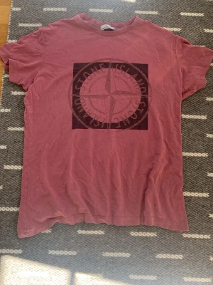 Rosa Stone Island t-shirt med tryck - Säljer en rosa t-shirt från Stone Island med stort ikoniskt kompass-tryck på bröstet. T-shirten har klassisk passform och rund hals. Perfekt för dig som gillar streetwear och vill sticka ut lite extra.