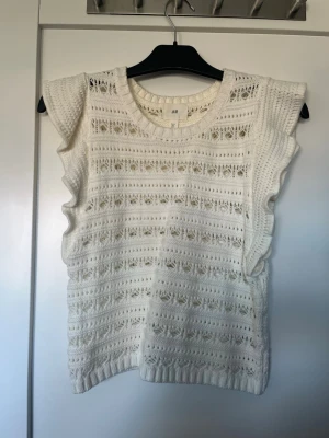 Vit ärmlös stickad topp från H&M - Säljer en vit ärmlös stickad topp från H&M med snyggt hålmönster och ribbade kanter. Toppen har rund halsringning och volangdetaljer längs ärmhålen. Perfekt för lager på lager och ger en fräsch look till din outfit.