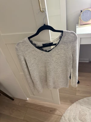 Ljusgrå tröja med spetsdetalj - Snygg ljusgrå tröja från Veromoda med v-ringning och svart spetskant runt halsen. Tröjan har en rak passform och långa ärmar, perfekt för lager-på-lager-stil. Mjuk och skön att bära, passar till många olika outfits.