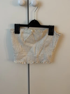 Vit bandeau tubtopp - Säljer en söt vit bandeau tubtopp från BikBok i smockat tyg. Toppen är ärmlös och har volangkant upptill och nedtill för en extra gullig touch. Perfekt att matcha med höga jeans eller kjol. Gåva vid köp!