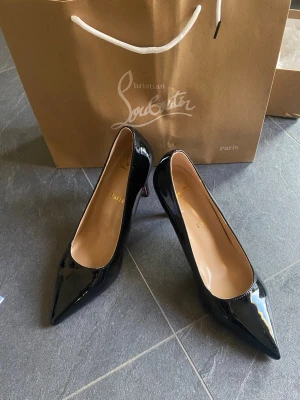 Svarta lackpumps från Christian Louboutin - Snygga svarta pumps i lack från Christian Louboutin med klassisk spetsig tå och ikonisk röd sula. Skorna har hög klack och elegant form, perfekta för att lyfta vilken outfit som helst. Materialet är glansigt lackläder och insidan är beige.
