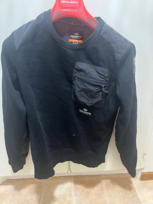 Parajumpers tröja - Cool svart sweatshirt från Parajumpers med en stor ficka med dragkedja på bröstet och patch med logga på ärmen. Tröjan har rund halsringning och ribbade muddar. Perfekt för en streetwear-look.