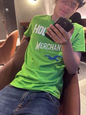 Grön Hollister t-shirt med tryck - Snygg grön t-shirt från Hollister med vit text och blå logga på bröstet. Klassisk passform och rund halsringning. Perfekt till jeans eller shorts för en avslappnad look.