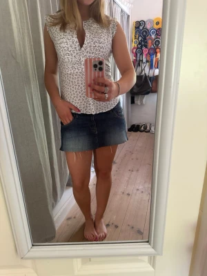 Vit spetsblus med detaljer - Söt vit blus med snkare och liten ståkrage. Ärmlös modell med lätt volang vid axlarna och en liten v-ringning framtill. Perfekt till jeans eller kjol för en fräsch och somrig look. Materialet känns luftigt och mjukt.