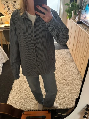 Randig jeansskjorta i blått och vitt - Säljer en jeansskjorta med blåa och vita ränder. Skjortan har knäppning framtill, bröstficka och klassisk krage. Använd men inga tydliga defekter.