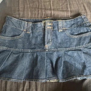 Blå jeanskjol med volangkant och inbyggda shorts - Trendig blå jeanskjol med volangkant och dubbla knappar framtill. Kjolen har klassiska fickor fram och bak, bälteshällor, inbyggda shorts och kontrastsömmar. Perfekt för dig som gillar en avslappnad men snygg stil.